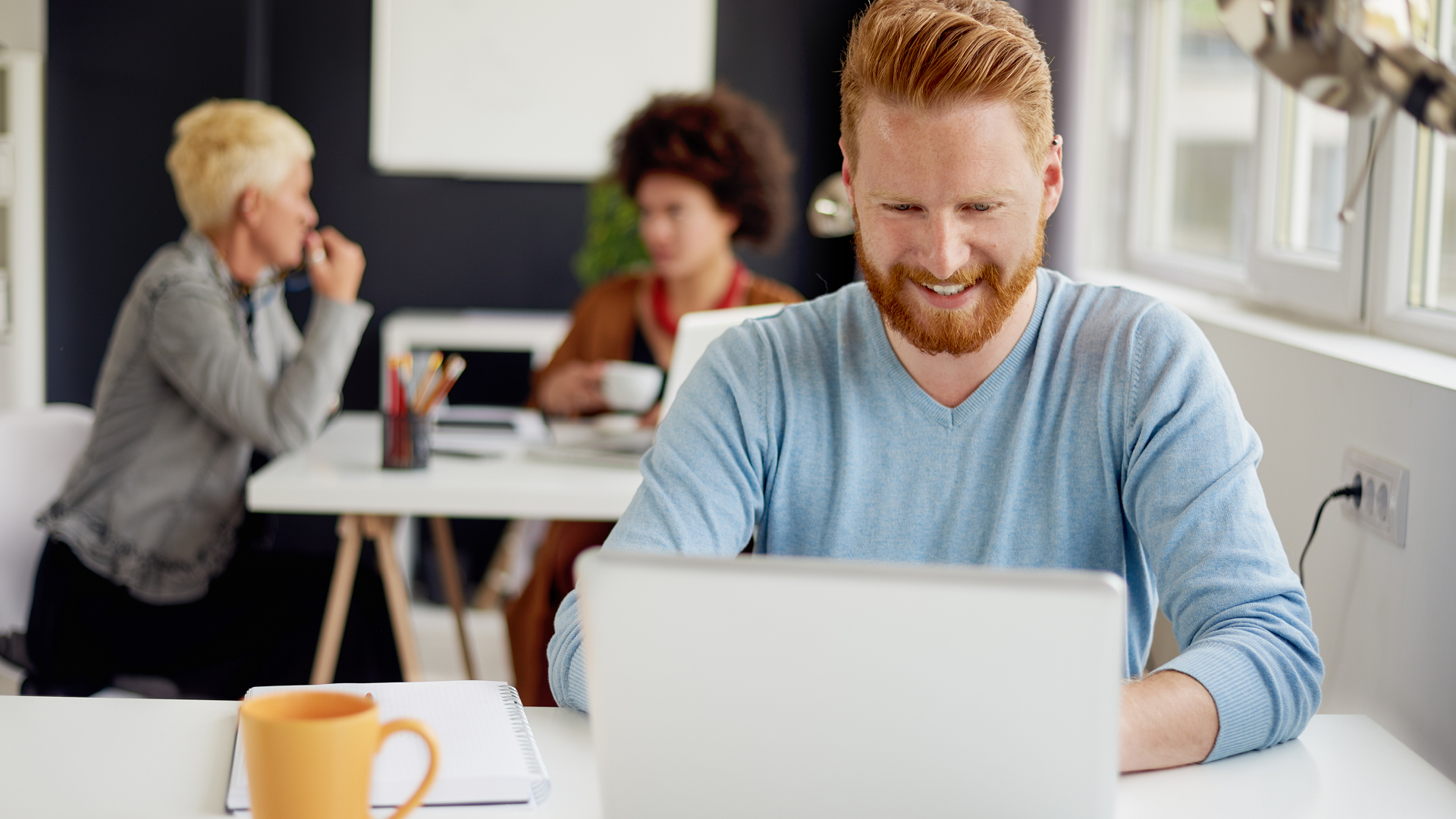 Businessman on laptop smiling at screen with coworkers in background.