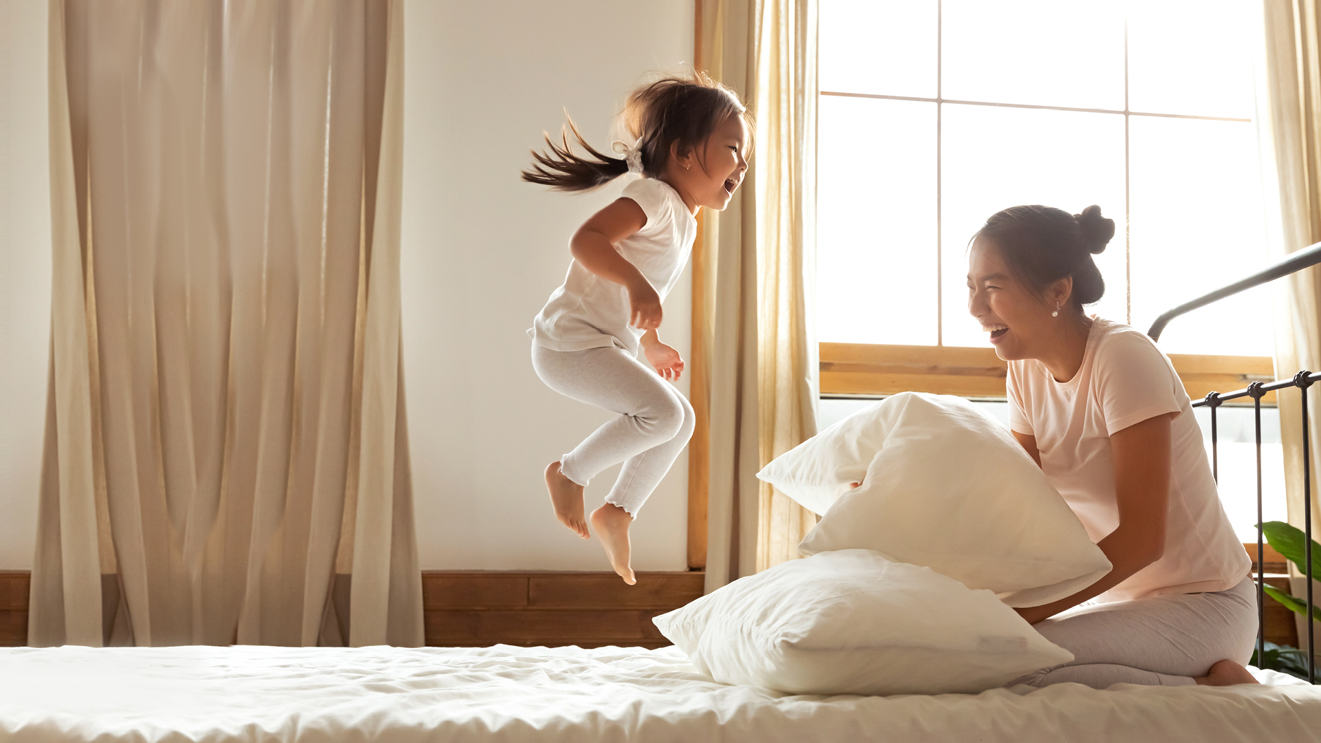 Mother laughs as daughter jumps on bed.